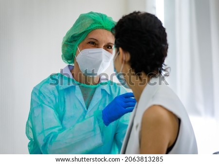 Caucasian senior female doctor in PPE full hazard protection uniform with face mask using syringe needle inject vaccine to woman patient shoulder at hospital vaccinating desk.