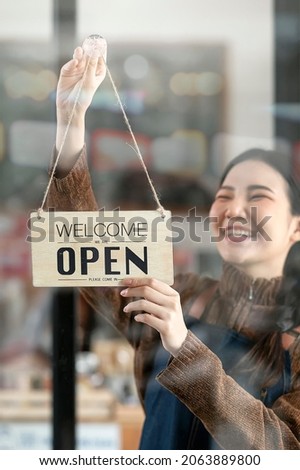 Young smiley female entrepreneur holding open sign board hanging on window glass of cafe.