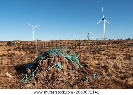 Stack of turf in a bog next to wind farm turbine. Clean new and dirty old source of energy. Natural and artificial product in nature environment. Warm sunny day. Nobody