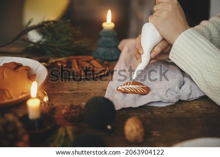 Hands decorating gingerbread cookie christmas tree with frosting on rustic table with napkin, candle, spices, decorations. Atmospheric moody image. Making traditional christmas gingerbread cookies