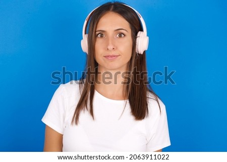 Serious displeased Young european woman wearing white T-shirt on blue background looks puzzled at camera being angry wears stereo headphones listens music while walking at street