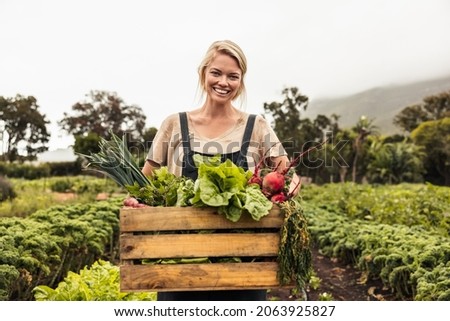 Cheerful organic farmer holding a box full of fresh produce on her farm. Happy young woman smiling at the camera while standing in her vegetable garden. Successful female farmer harvesting vegetables. Royalty-Free Stock Photo #2063925827