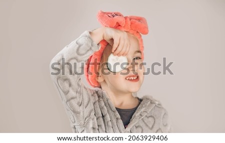 A little girl wearing bathrobe, hair band and holding pieces of cucumber isolated over beige background