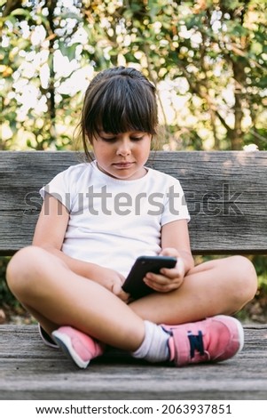 Little black-haired girl, wearing a white t-shirt, sitting on a bench in a park, looking at the mobile phone, smiling.