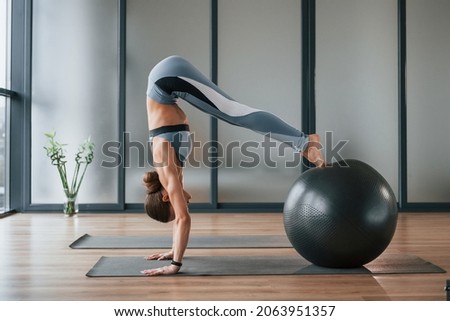 Uses fitness ball. Young woman in sportive wear and with slim body have yoga day indoors.