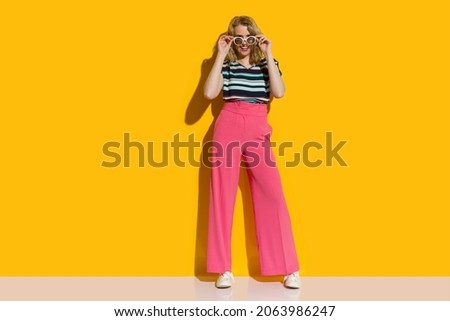 Beautiful young woman in sunglasses, high waisted pink baggy pants and striped top is posing in front of yellow wall. Front view. Full length shot.