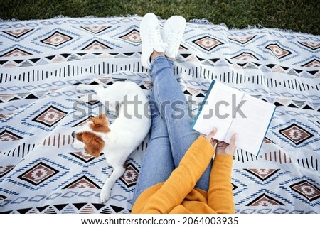 top view of woman having fun with jack russell dog in park, sitting on blanket during autumn season. Woman reading a book while cuddling dog. Pets and love concept