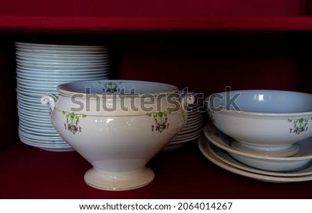 Antique service of Italian ceramic plates with a dark red background. Dinner plates and tureen on a fabric-covered shelf. Stacks of ceramic retro plates. Vintage image. Royalty-Free Stock Photo #2064014267