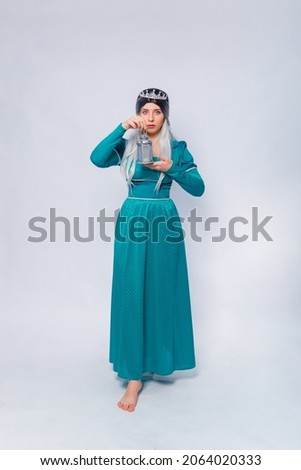 Full length portrait of a princess in a medieval, fantasy, turquoise dress with ash hair and a silver crown, posing with a lamp in her hands isolated on a white background.

