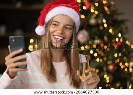 Happy girl in Santa hat posing for selfie with perfect toothy smile, holding festive wine glass at Christmas tree, taking picture on smartphone, talking on video call. New Year distance celebration