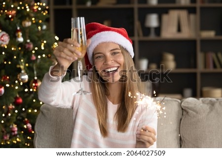 Happy joyful Xmas girl in Santa hat toasting wineglass, looking at camera, smiling. Woman celebrating New year at home, drinking wine, holding burning sparklers on couch at Christmas tree