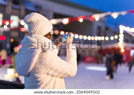 A woman on the rink is skating and taking selfie on smartphone. New Years Eve and Christmas. Fairy lights. Ice and snow mood concept. Winter sport.