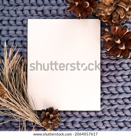 Top view photo of winter composition on purple knitting background with white blanc sheet of paper, branch and pine cones. Warm magic atmosphere. Greeting card, mockup for illustration, design