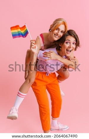 White lesbian couple piggybacking with rainbow flag isolated over pink background Royalty-Free Stock Photo #2064067490
