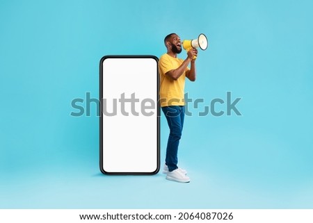 Happy young black guy shouting into megaphone, standing near big smartphone with empty screen, promoting your mobile app or website, offering mockup space for advertisement, blue background Royalty-Free Stock Photo #2064087026