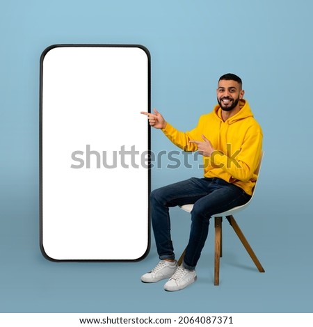 Positive arab guy pointing at big smartphone with white screen while sitting on chair over blue background. Man demonstrating free space for your app or website design, mockup Royalty-Free Stock Photo #2064087371