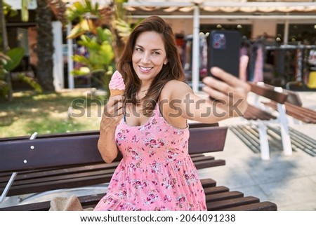 Brunette woman enjoying a summer day sitting on a bench at the park eating a ice cream cone taking a selfie picture
