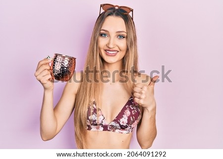 Young blonde girl wearing bikini drinking cocktail smiling happy and positive, thumb up doing excellent and approval sign 