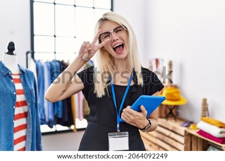Young blonde woman working as manager at retail boutique doing peace symbol with fingers over face, smiling cheerful showing victory 