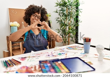 Beautiful african american woman with afro hair painting at art studio smiling in love doing heart symbol shape with hands. romantic concept. 
