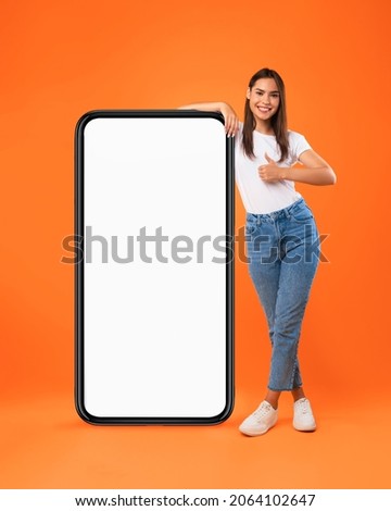 Mobile Offer. Happy woman leaning on huge cellphone with blank white screen, showing thumb up gesture, recommending great new app or website for phone, offering space for ad, mock up, full body length Royalty-Free Stock Photo #2064102647