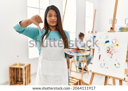 Young latin woman standing at art studio with angry face, negative sign showing dislike with thumbs down, rejection concept 