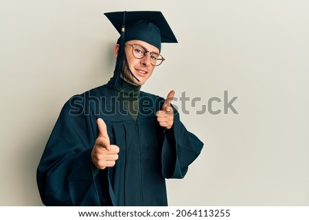 Young caucasian man wearing graduation cap and ceremony robe pointing fingers to camera with happy and funny face. good energy and vibes. 