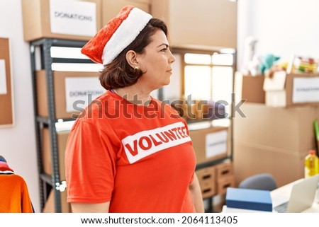 Middle age hispanic woman wearing volunteer t shirt and christmas hat looking to side, relax profile pose with natural face with confident smile. 