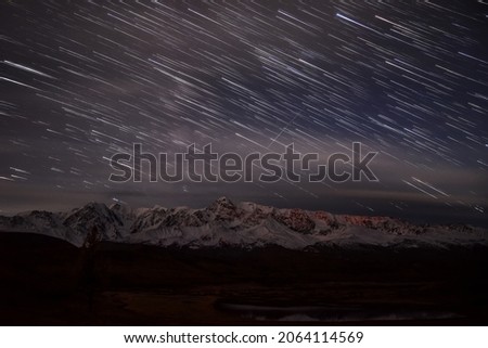 Amazing night view with stars in the form of tracks and blurry clouds in the sky over snowy mountains, forest and lake in moonlight in autumn. Altai, Russia