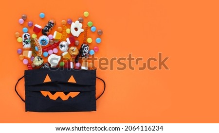 Black medical mask like smiling pumpkin jack-o-lantern gift bag full of spilled assorted traditional Halloween candies. Orange banner background with copy space. Protection from Covid-19 concept. 