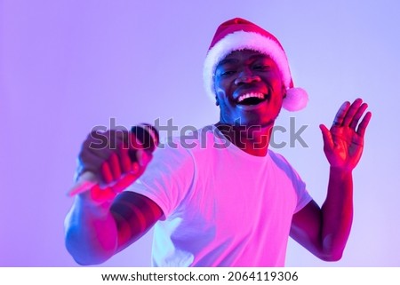Christmas music. Cool black guy in Santa hat singing holiday song, using microphone, performing karaoke in neon light. Carefree African American singer giving live music concert on New Years eve