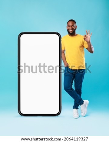 Best mobile offer. Smiling black guy demonstrating giant smartphone with screen mockup and showing okay gesture on blue studio background, recommending your app or website