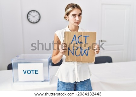 Young blonde woman at political election holding act now banner thinking attitude and sober expression looking self confident 