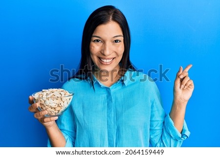Young latin woman holding bowl with pumpkin seeds smiling happy pointing with hand and finger to the side 