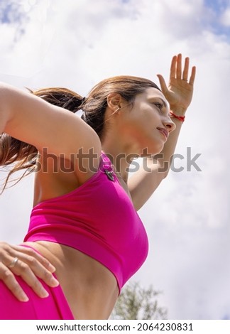 Young woman practicing pilates and stretching in a park in a cloudy day on summer. Long haired caucasian woman in a pink top and leggins doing sport outdoors. Healthy lifestyle concept. Below view Royalty-Free Stock Photo #2064230831