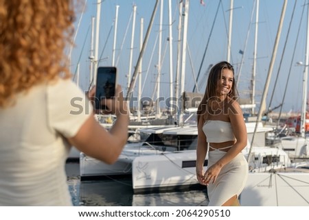 a red-haired girl with curly hair taking a picture of her long-haired brunette friend in a white dress and laughing on the pier with the boats and the ocean
