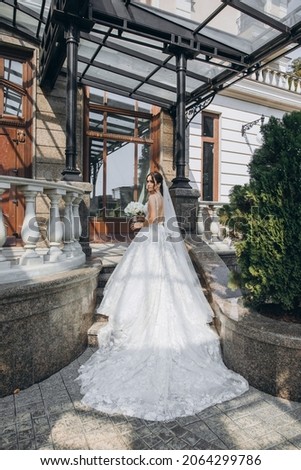 beautiful bride stands in a beautiful dress with a bouquet in her hands near a large gray house and looks to the camera