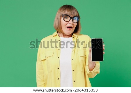 Elderly shocked happy woman 50s in glasses yellow shirt hold in hand use mobile cell phone with blank screen workspace area isolated on plain green background studio portrait People lifestyle concept