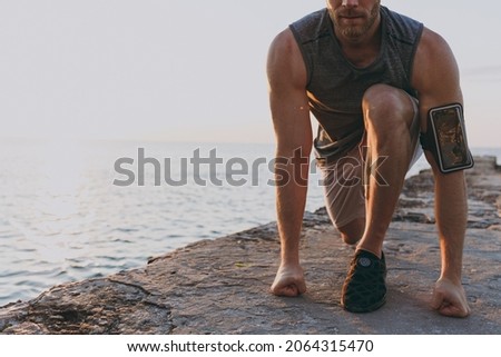 Cropped photo young strong toned fit sportsman man 20s wearing sport clothes mobile phone warm up train start sprint at sunrise sun dawn over sea beach outdoor on pier seaside in summer day morning