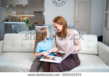 mother and daughter at home on the couch leafing through and looking a book with photos from a family photo shoot with a newborn baby. memory of important moments of life in the photo album. Royalty-Free Stock Photo #2064333293