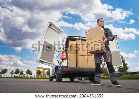postman next to van. Mail van is in background. Courier to postal service. Postman in gray uniform. Courier took box out of van. Employee of postal delivery service. Career in courier company Royalty-Free Stock Photo #2064351359