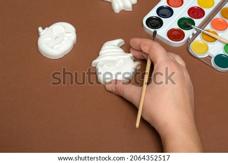 Halloween crafts. Gibs figurines of pumpkins and ghosts with bright paint with a brush on a brown background, ready for drawing close-up. A woman draws with a calligraphic brush on a white pumpkin. 
