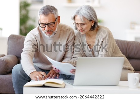 Worried senior family couple reading financial documents or notification letter with bad news from bank while paying bills online on laptop, retired husband and wife having money problems