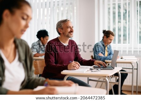 Happy mature student writing notes and using laptop while learning during a class at lecture hall. Royalty-Free Stock Photo #2064472385
