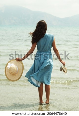 Image of a young brunette woman in a blue dress walking barefoot on a beach and dangles his feet in the water. Young woman in summer white dress with straw hat looking to a sky and sea. Back view. Royalty-Free Stock Photo #2064478640