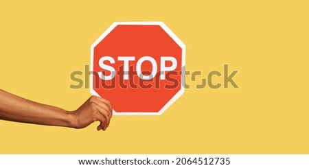 Closeup of male hand holding octagonal red Stop symbol, ban concept, arm showing traffic sign, prohibition concept, forbidden way, no access. Indoor studio shot isolated on yellow background. Royalty-Free Stock Photo #2064512735