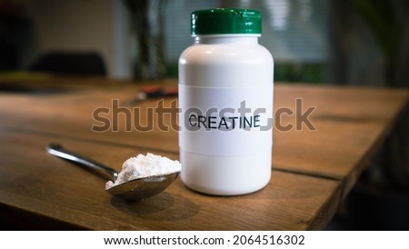 Close shot of white plastic bottle with the word Creatine written on the label, with a silver spoon of white powder at the side set on a wooden table with an out of focus background. Royalty-Free Stock Photo #2064516302