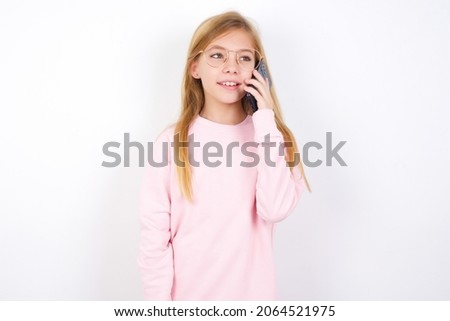 Pleasant looking happy beautiful caucasian little girl with glasses wearing pink sweatshirt on white background has nice telephone conversation and looks aside, has nice mood and smiles positively 