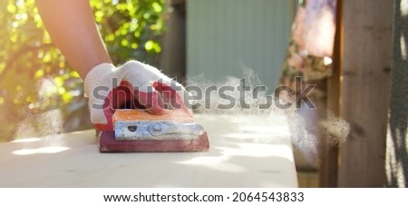 A man polishes a wooden surface with sandpaper. A male hand in protective gloves works outdoors. Sawdust dust glows in the sun. Horizontal format, close-up, side view Layout with space for text. Royalty-Free Stock Photo #2064543833