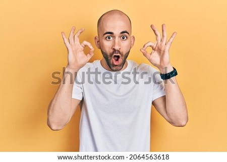 Young bald man wearing casual white t shirt looking surprised and shocked doing ok approval symbol with fingers. crazy expression 
